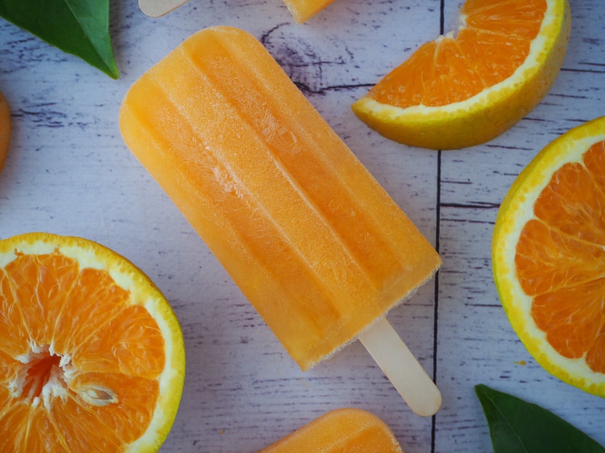 Orange popsicle on a diagonal, with fresh oranges and orange leaves.