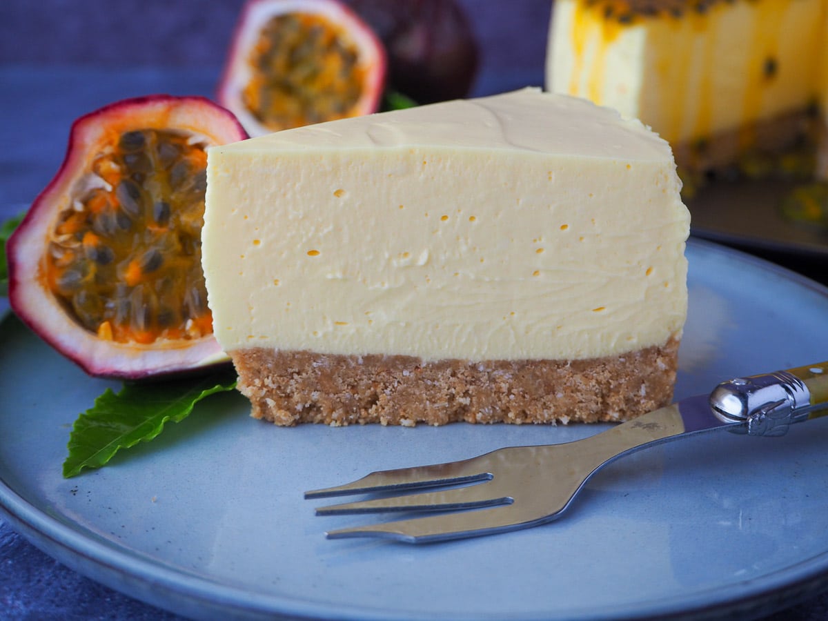 Passion fruit cheesecake on a plate, with a vintage for, fresh passion fruit and leave on the side, and the rest of the cake in the background.