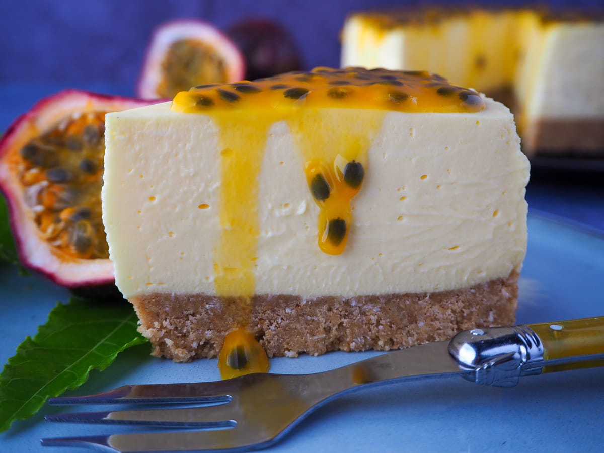 Passion fruit cheesecake with passion fruit coulis, a vintage fork in front, fresh passion fruit and passion fruit leave on the side, and rest of cheesecake in the background