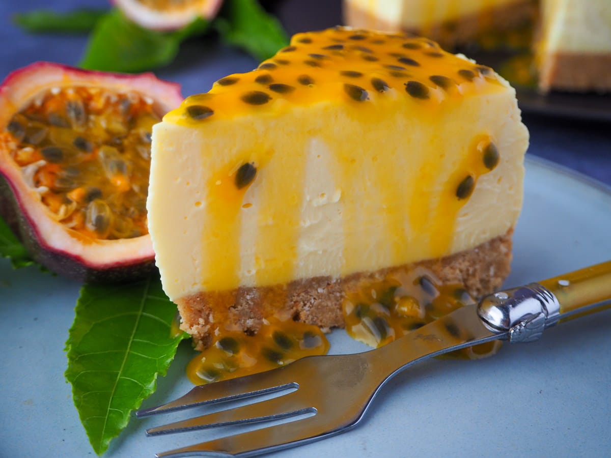 Passion fruit cheesecake with passion fruit coulis, a vintage fork in front, fresh passion fruit and passion fruit leave on the side, and rest of cheesecake in the background