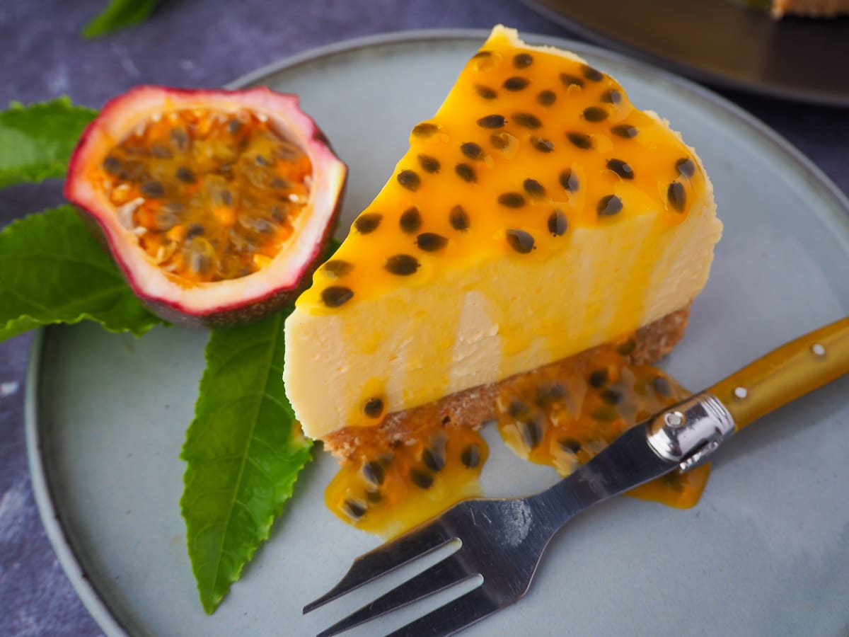 Passion fruit cheesecake with passion fruit coulis, a vintage fork in front, fresh passion fruit and passion fruit leave on the side, and rest of cheesecake in the background