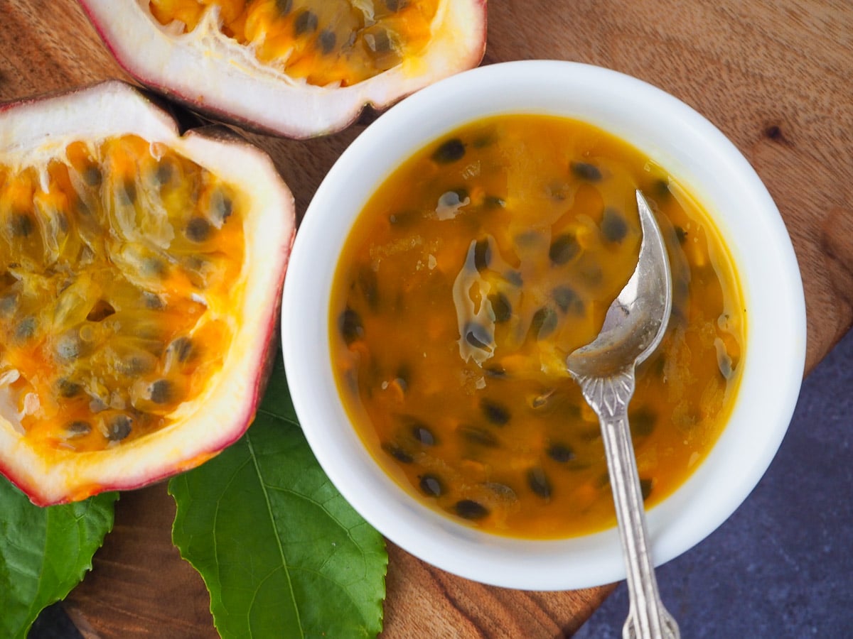 Passion fruit coulis a small white bowl with a vintage spoon in it and fresh, cut open passion fruit and passion fruit leaves on the side, on a wooden chopping board.