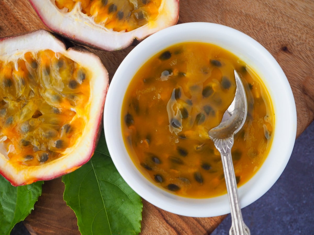 Passion fruit coulis 3 Passion fruit coulis a small white bowl with a vintage spoon in it and fresh, cut open passion fruit and passion fruit leaves on the side, on a wooden chopping board.