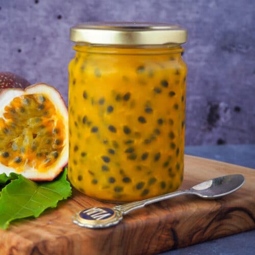 Passion fruit coulis in a glass jar with a vintage spoon in front and fresh, cut open passion fruit and passion fruit leaves on the side, on a wooden chopping board.