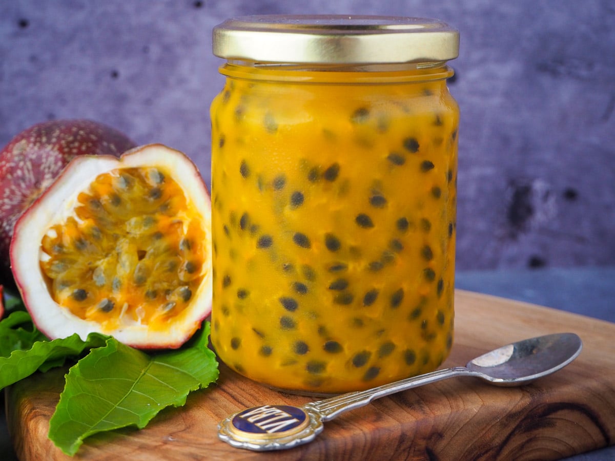 Passion fruit coulis in a glass jar with a vintage spoon in front and fresh, cut open passion fruit and passion fruit leaves on the side, on a wooden chopping board.