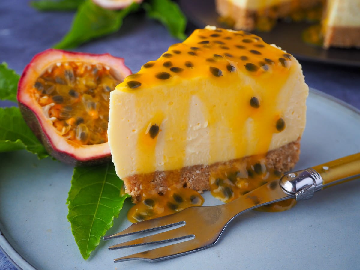 Passion fruit coulis over slice of cheesecake, with a vintage fork, fresh passion fruit and leaves on the side, and rest of the cheesecake in the background.