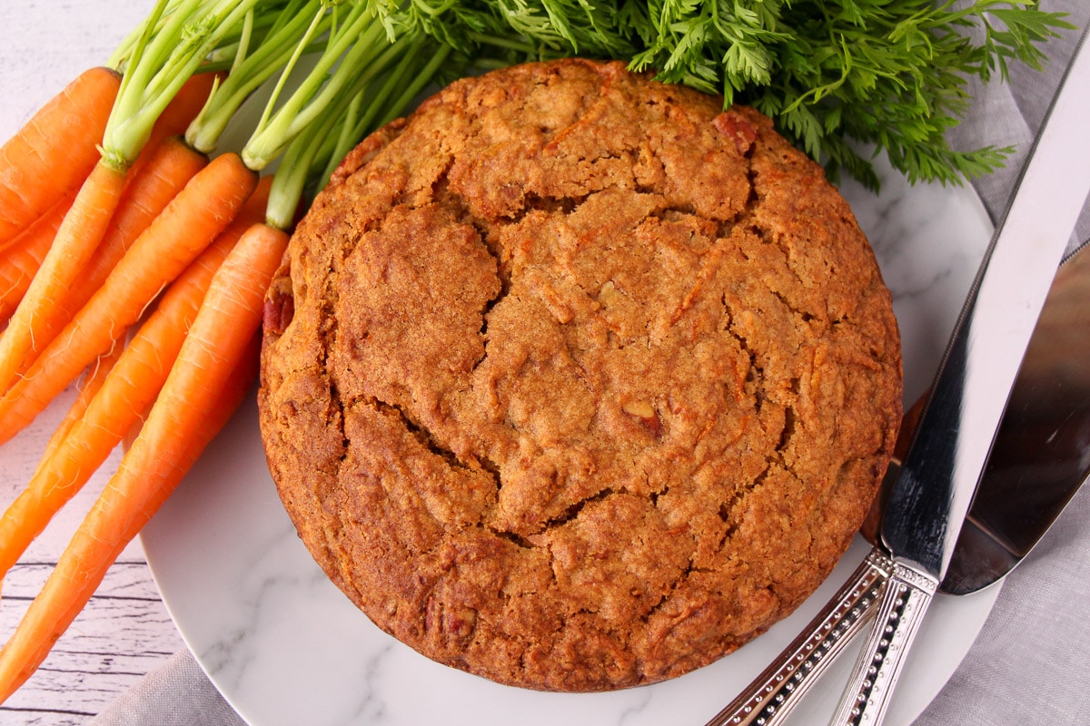 Top down shot of whole un decorated eggless carrot cake, with fresh Dutch carrots on one side and silver serving wear on the other.