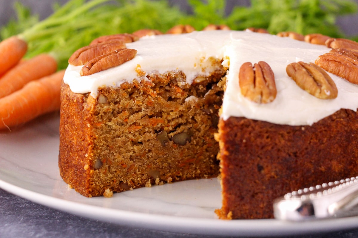 Eggless carrot cake decorated with lemon icing and pecans, with single slice taken out, fresh Dutch carrots and silver serving wear in the background.