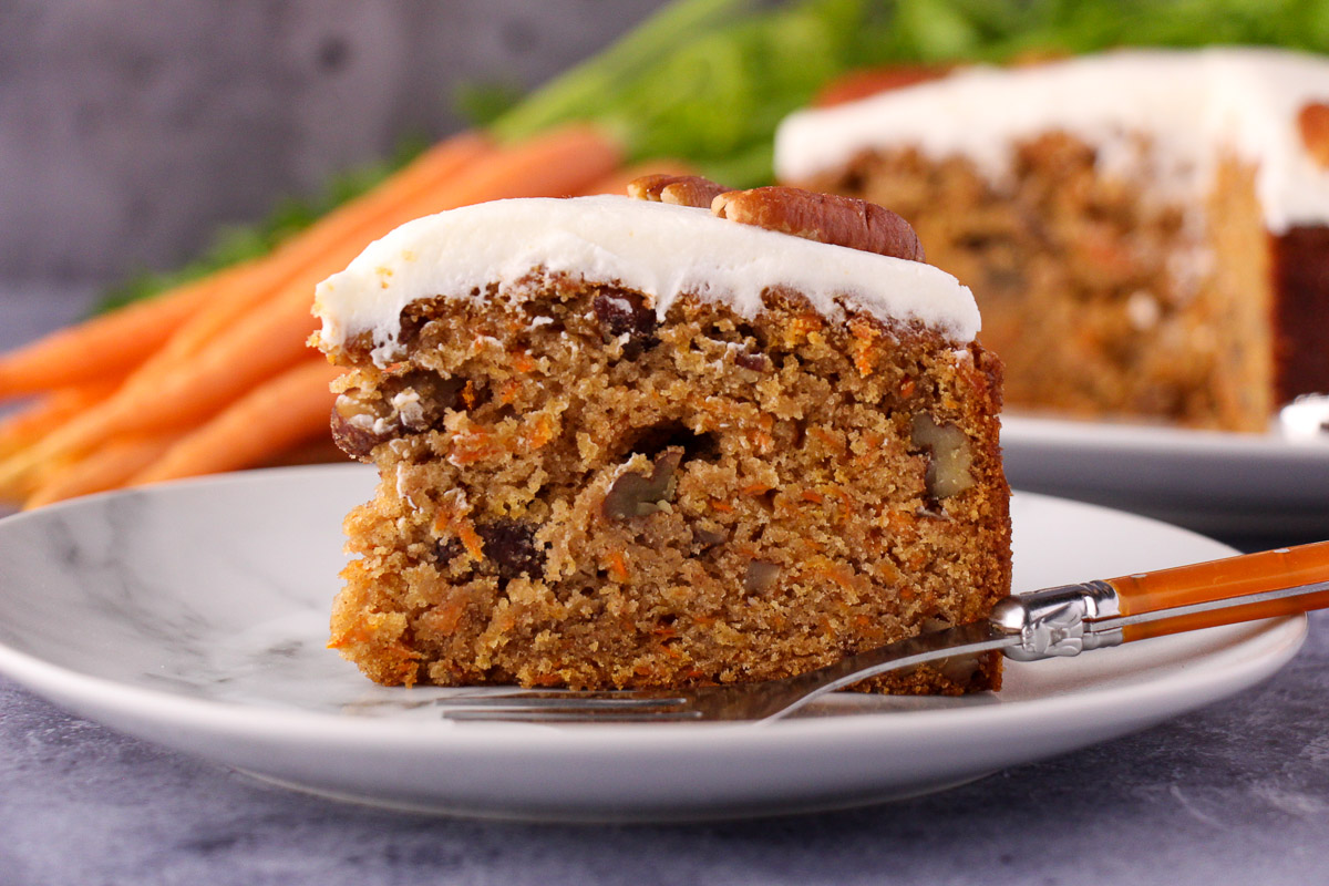 Slice of eggless carrot cake decorated with lemon icing and pecans on a plate with a vintage fork and the rest of the cake and fresh Dutch carrots in the background.