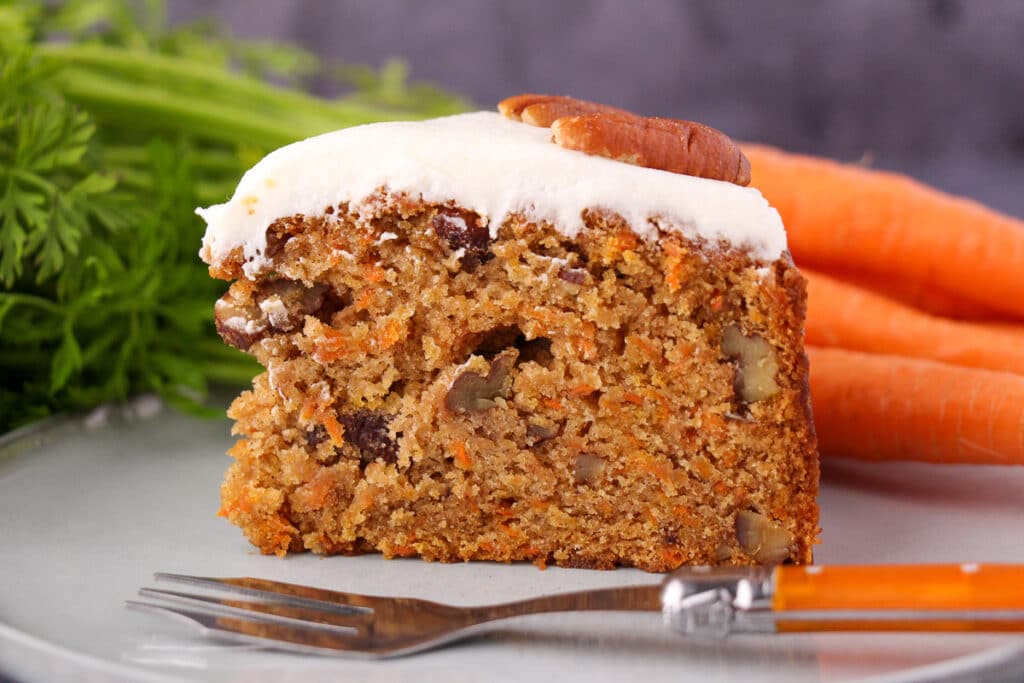 Slice of eggless carrot cake decorated with lemon frosting and pecans, on a plate with a vintage fork and fresh Dutch carrots in the background.