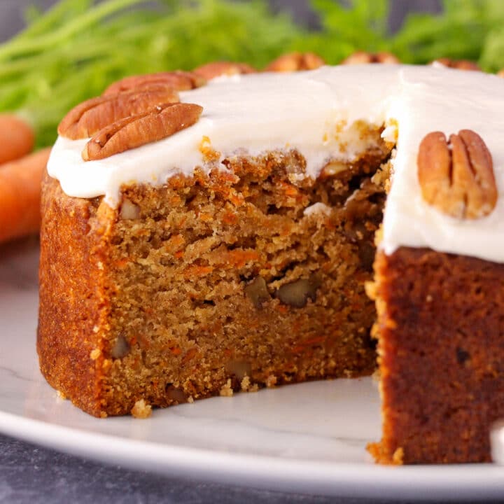 Close up eggless carrot cake decorated with lemon frosting and pecan, with fresh Dutch carrots in the background.