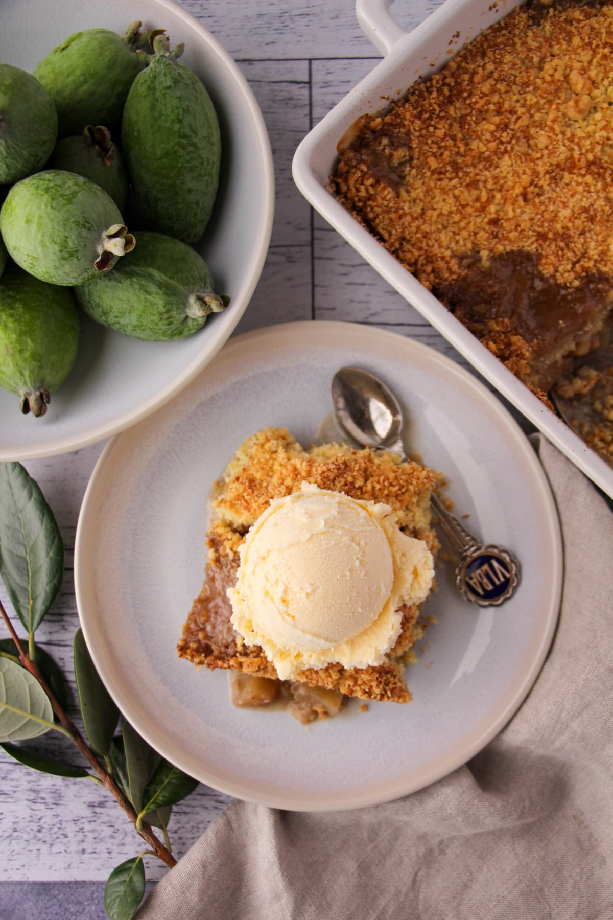 Feijoa crumble on a plate with a scoop of ice cream on top, vintage spoon on the side and fresh feijoas and baking dish of crumble on the sides.