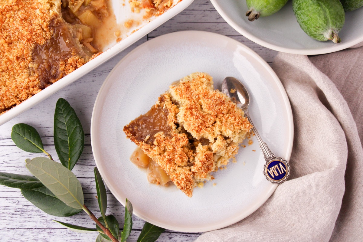 Feijoa crumble on a plate with a vintage spoon on the side and fresh feijoas and baking dish of crumble on the sides.