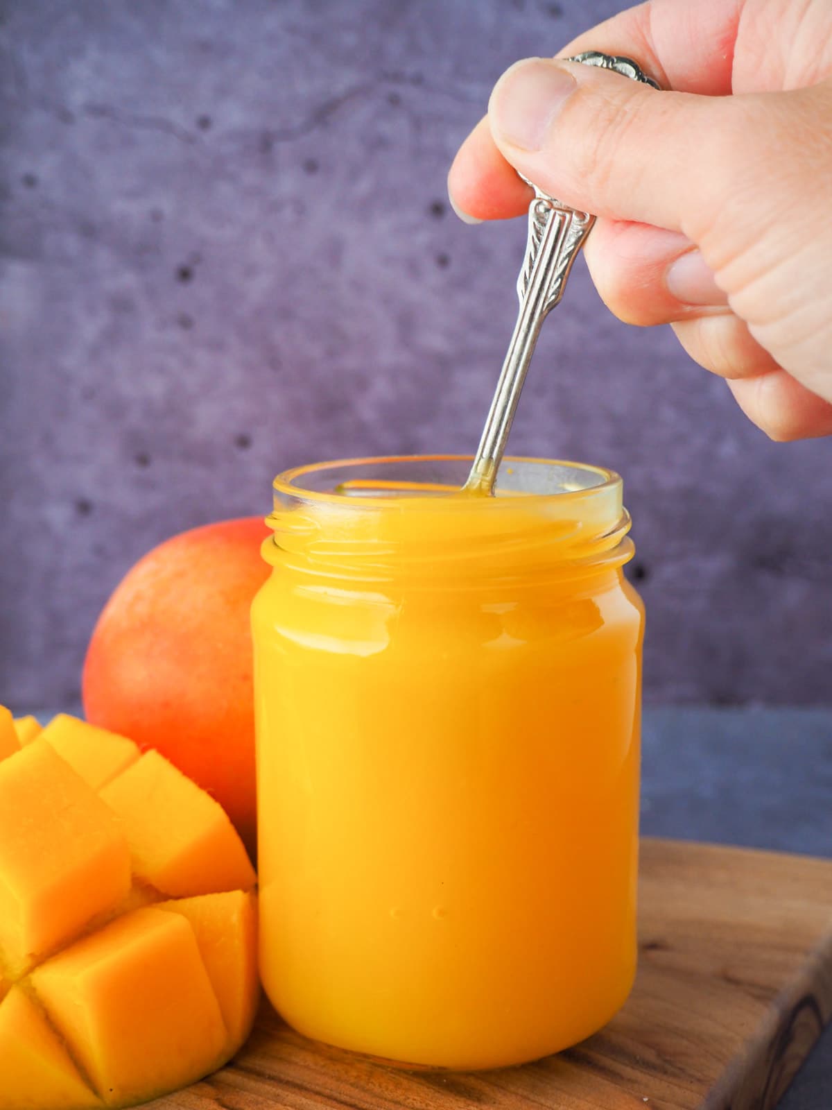 Glass jar of mango coulis with a hand holding a spoon in the jar, with fresh mango on the side.