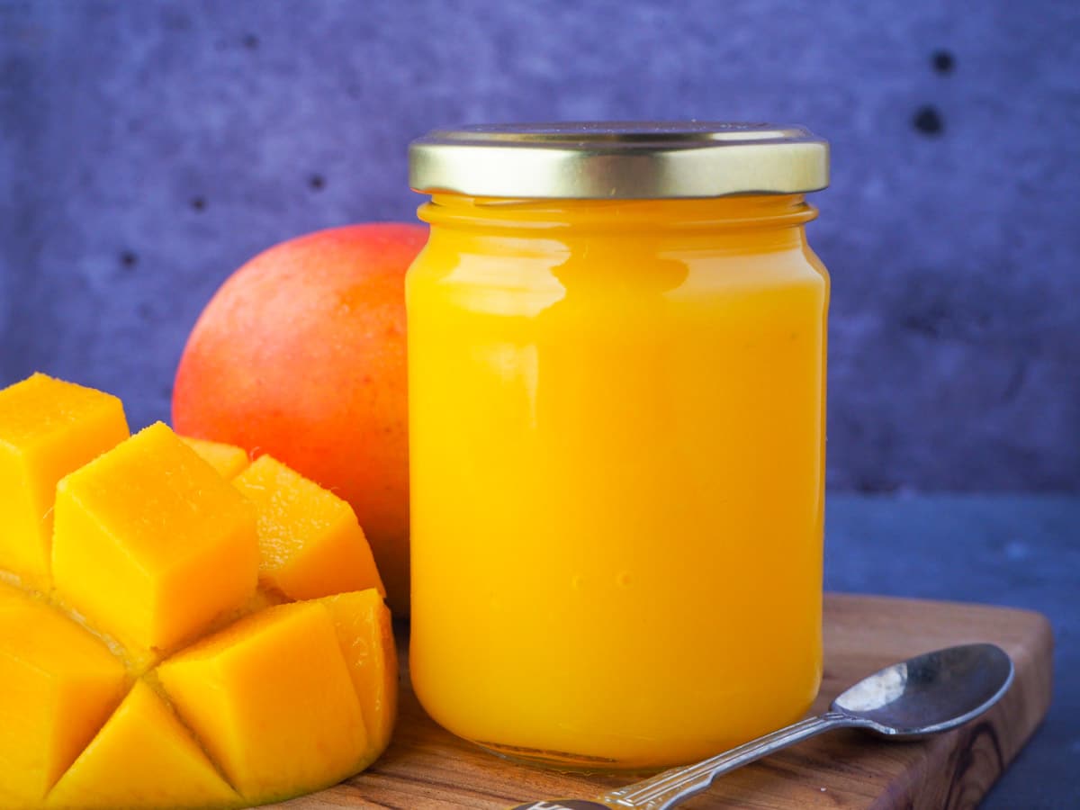 Glass jar of mango coulis with fresh mango and a vintage spoon on the side.