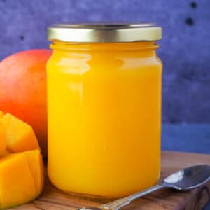Close up closed glass jar of mango coulis with fresh mango and a vintage spoon on the side.