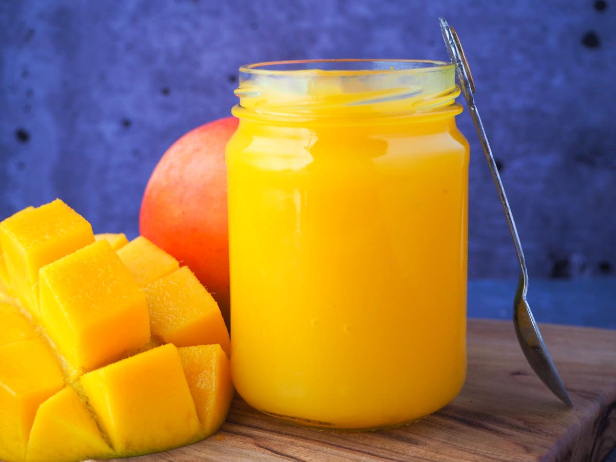 Open glass jar of mango coulis with fresh mango and a vintage spoon propped on the side.