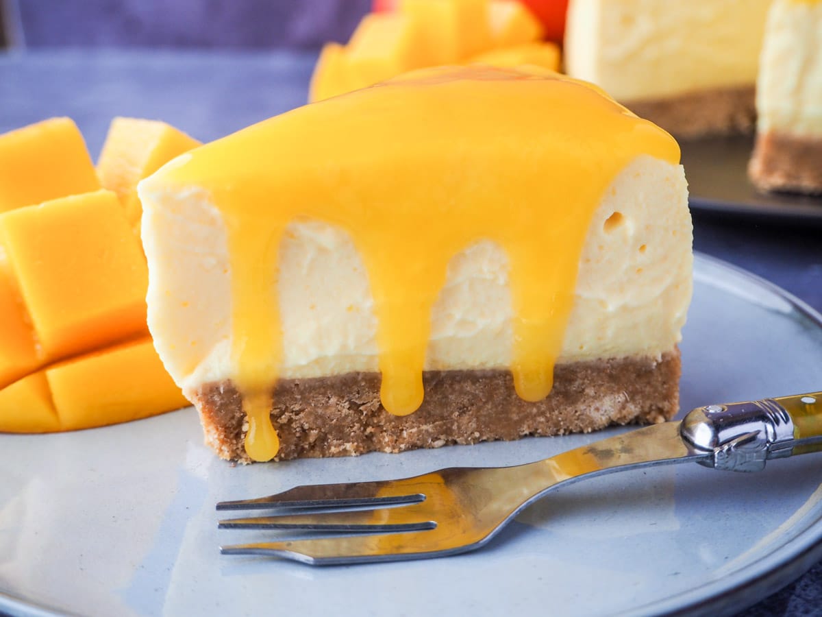 Slice of mango cheesecake with mango coulis oozing over the sides, on a plate with a vintage fork, fresh mango and the rest of the cheesecake in the background.