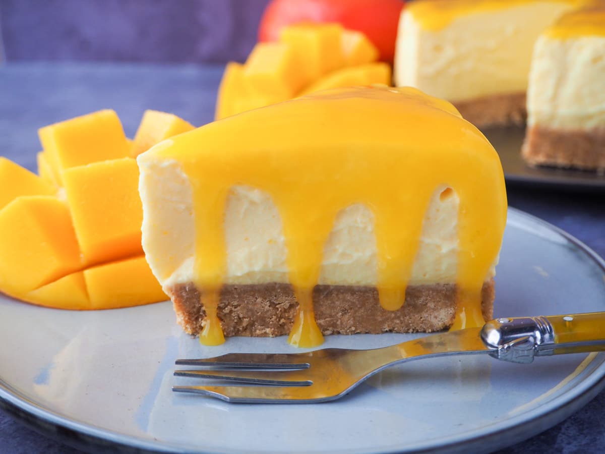 Slice of no bake mango cheesecake with mango coulis, a vintage fork and fresh mango on the side.