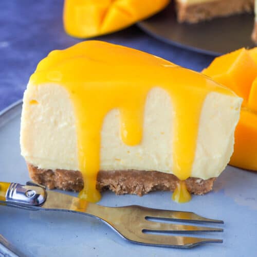 Close up slice of no bake mango cheesecake with mango coulis, a vintage fork and fresh mango on the side.
