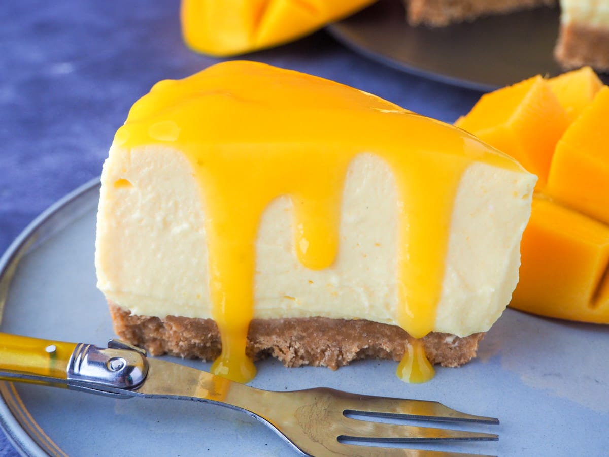 Slice of no bake mango cheesecake with mango coulis, a vintage fork and fresh mango on the side.