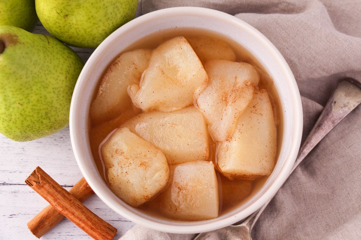 Stewed pears in a bowl, with fresh pears, cinnamon sticks and a vintage serving spoon on the side.