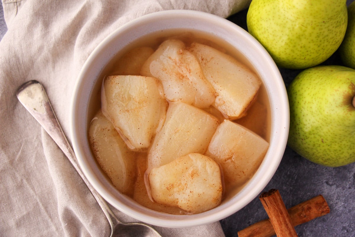 Stewed pears in a bowl, with fresh pears, cinnamon sticks and a vintage serving spoon on the side,