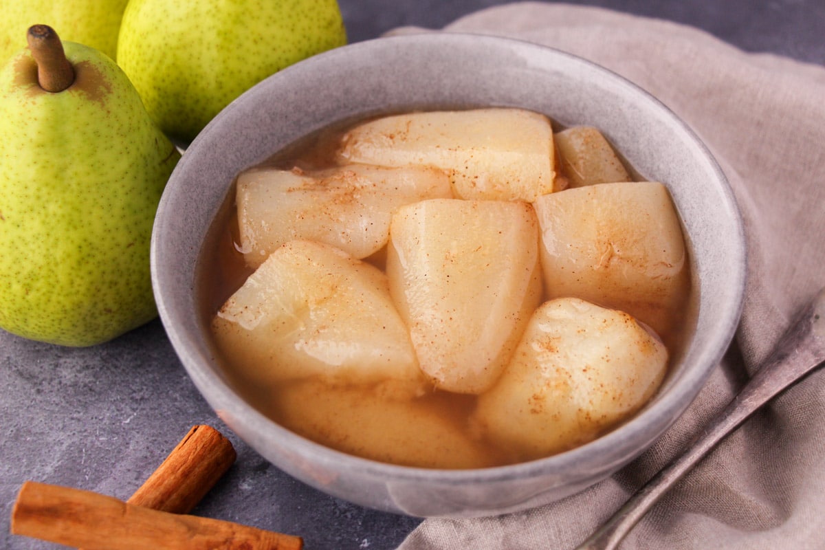 Stewed pears in a bowl, with fresh pears, cinnamon sticks and a vintage serving spoon on the side.