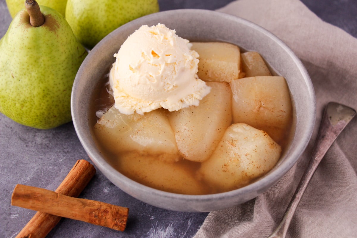 Stewed pears in a bowl with a scoop of vanilla ice cream, with fresh pears, cinnamon sticks and a vintage serving spoon on the side.