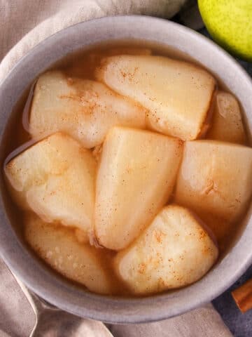 Close up stewed pears in a bowl, with fresh pears, cinnamon sticks and a vintage serving spoon on the side.