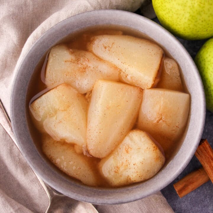 Close up stewed pears in a bowl, with fresh pears, cinnamon sticks and a vintage serving spoon on the side.