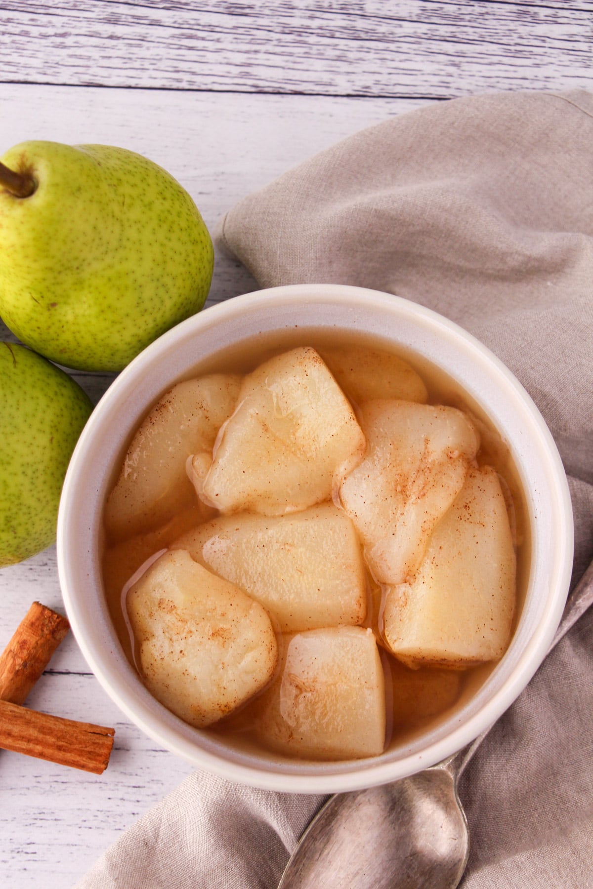 Stewed pears in a bowl, with fresh pears, cinnamon sticks and a vintage serving spoon on the side,