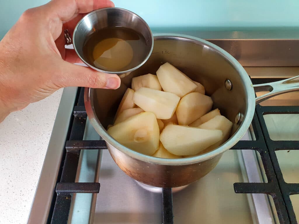 Adding water and vanilla essence mix to pears in pot on stove.