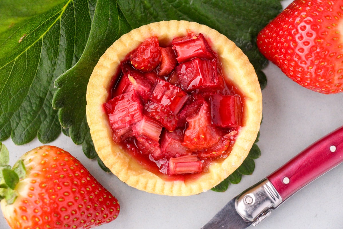 Close up strawberry rhubarb tartlets with strawberry leaves and fresh strawberries and a vintage cake fork on the side.