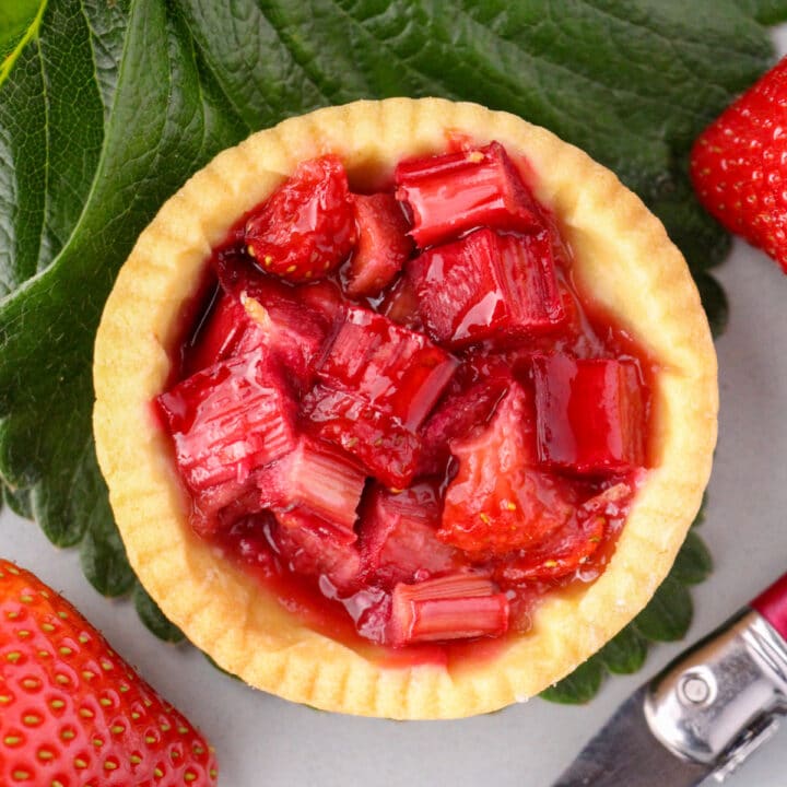 Close up strawberry rhubarb tartlets with strawberry leaves and fresh strawberries on the side.