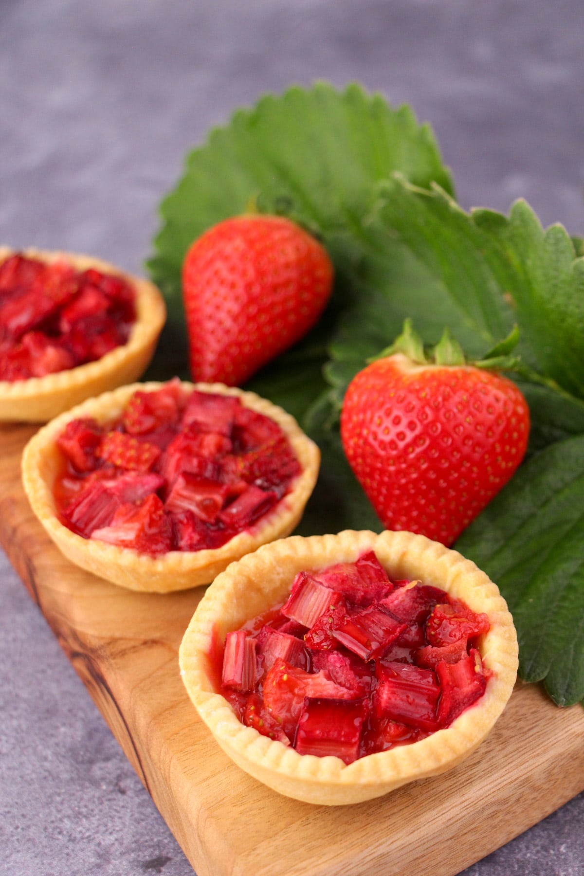 Row of strawberry rhubarb tartlets on a chopping board, with fresh strawberries and strawberry leaves on the side.