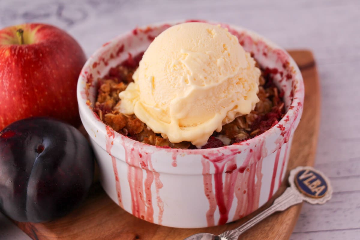 Apple and plum crumble in its ramekin with a scoop of vanilla ice cream and fresh fruit on the side.