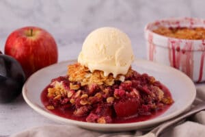 Apple and plum crumble on a plate with ice cream, with vintage serving spoon and ramakin and fresh fruit in the back ground