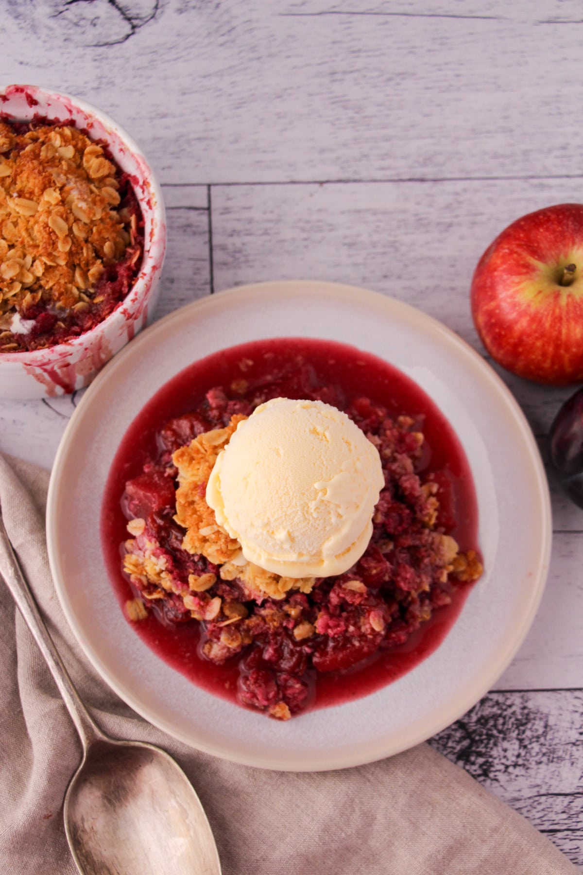 Apple and plum crumble on a plate with ice cream, with vintage serving spoon and ramakin and fresh fruit in the back ground.
