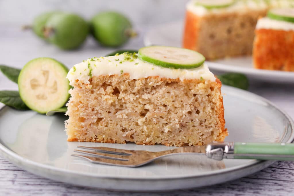 Slice of feijoa cake on a plate with a cake fork, the rest of the cake and fresh feijoas and leaves in the background.