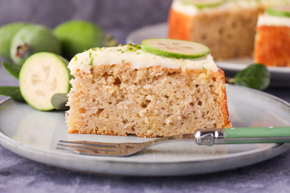 Slice of feijoa cake on a plate with a cake fork, the rest of the cake and fresh feijoas and leaves in the background.