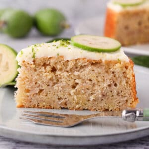 Close up slice of feijoa cake on a plate with a cake fork, the rest of the cake and fresh feijoas and leaves in the background.