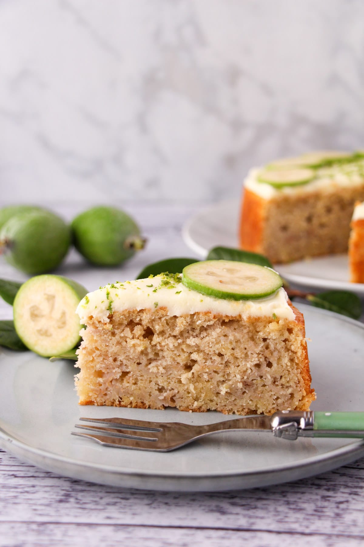 Slice of feijoa cake on a plate with a cake fork, the rest of the cake and fresh feijoas and leaves in the background.