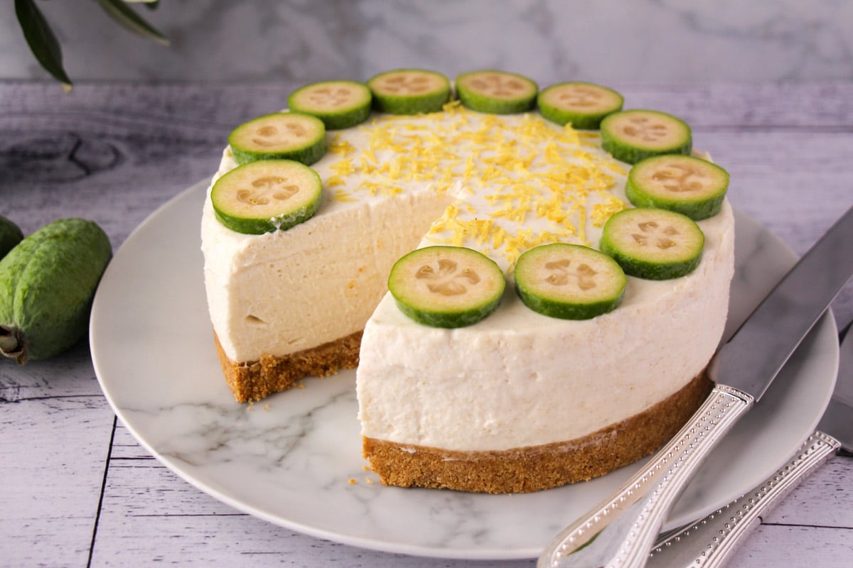 Feijoa cheesecake with slice taking out, silver serving ware and fresh feijoas and feijoa leaves in the background.