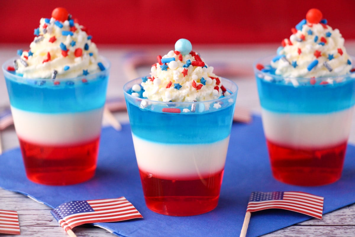 Red white and blue jello shots on blue napkin with whipped cream and sprinkles, surrounded by cocktail stick American flags.