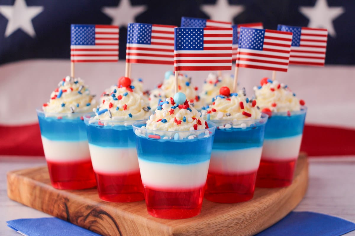 Red white and blue jello shots on a serving board with whipped cream, sprinkles, cocktail stick American flags with American flag in the background.