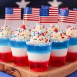 Close up red white and blue jello shots on a serving board with whipped cream, sprinkles, cocktail stick American flags with American flag in the background.