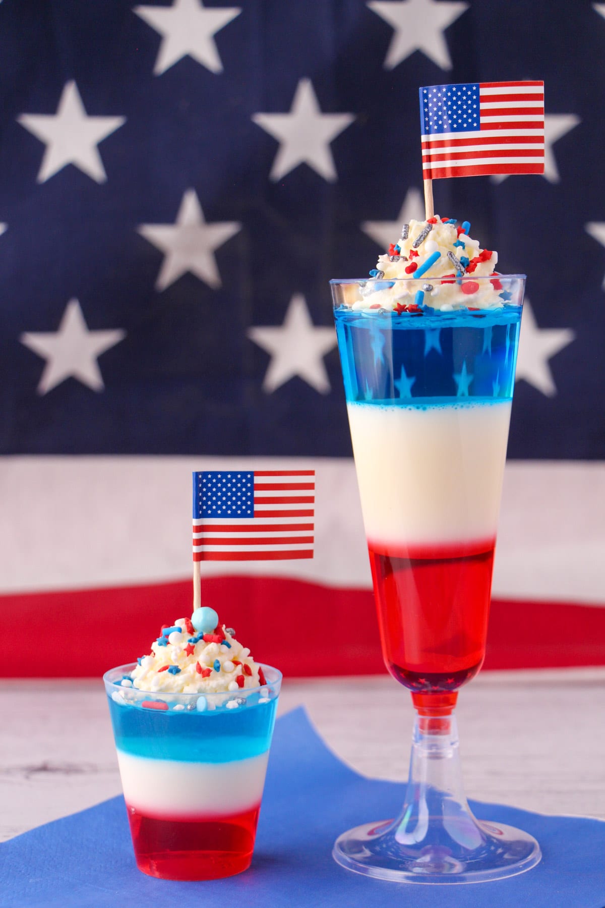 Kid friendly non alcoholic and alcoholic red white and blue jello shots in tall glasses with whipped cream, sprinkles, cocktail stick American flags with American flag in the background.