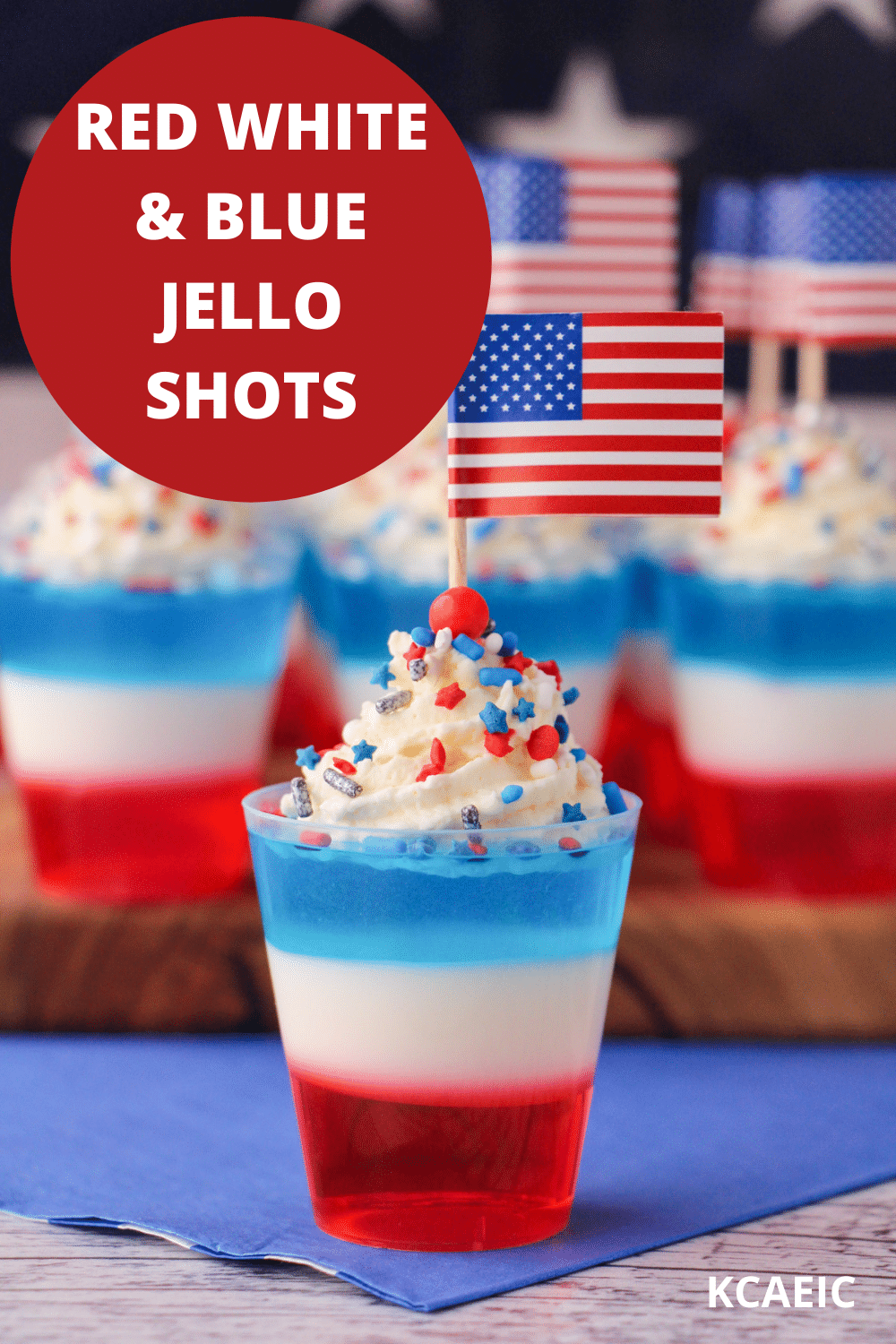 Red white and blue jello shots on a serving board with whipped cream, sprinkles, cocktail stick American flags with American flag in the background, with text overlay, red white and blue jello shots and KCAEIC.