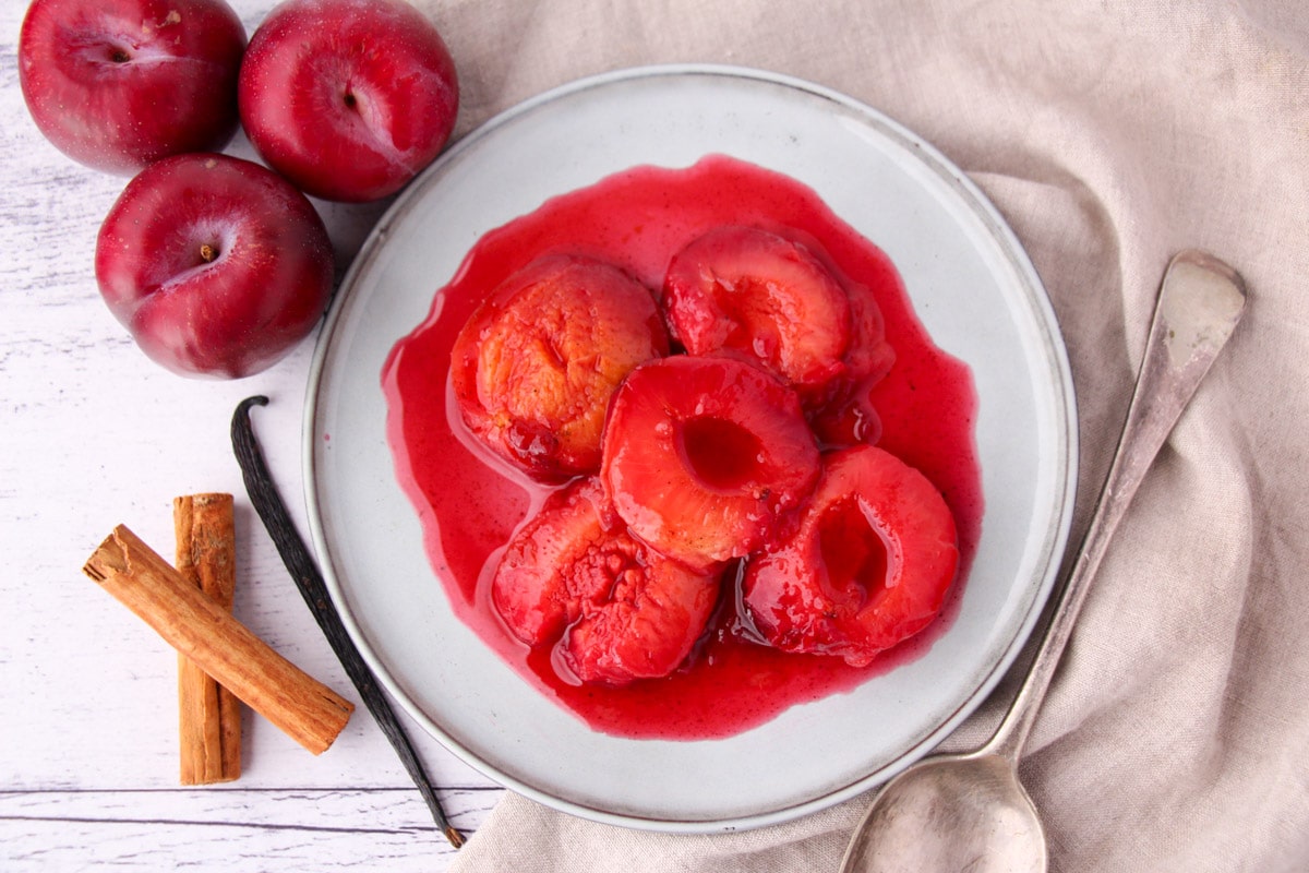 Stewed plums with a vintage serving spoon, fresh plums, cinnamon stick and vanilla pod on the side.